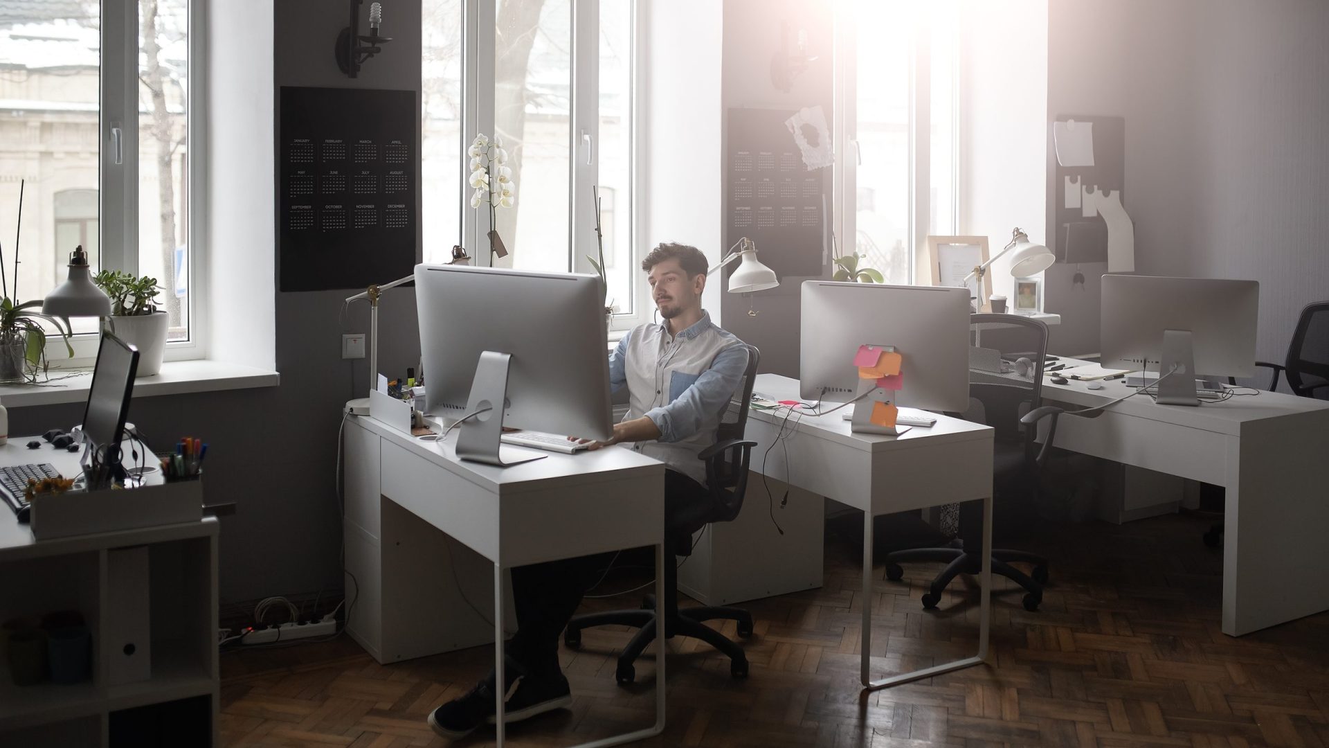Shot of young man standing at his desk and working on computer with light and windows in background.